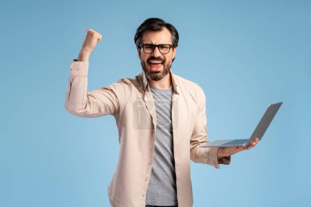 Photo for Happy bearded man in glasses rejoicing and feeling like champion, standing over blue background, holding laptop. Education, business, online technology concept - Royalty Free Image