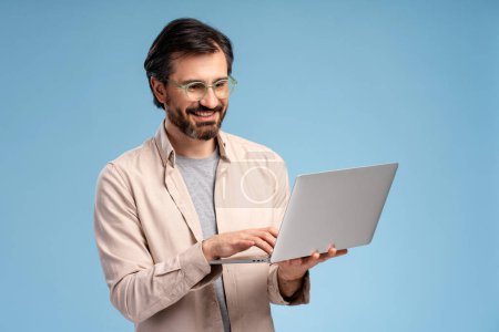Photo for Smiling positive bearded guy using laptop while typing, looking at screen, isolated on blue background. Education, business, online technology concept - Royalty Free Image