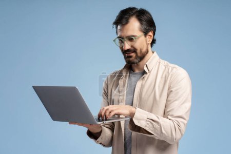 Photo for Busy young bearded man holding laptop and typing, looking at screen, posing in studio, isolated on blue. Smiling Education, business, online technology concept - Royalty Free Image