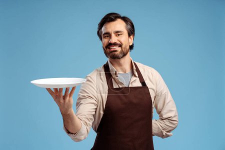 Photo for Portrait of happy bearded waiter in apron showing empty plate, looking at camera in studio, isolated on blue. Small business startup concept - Royalty Free Image