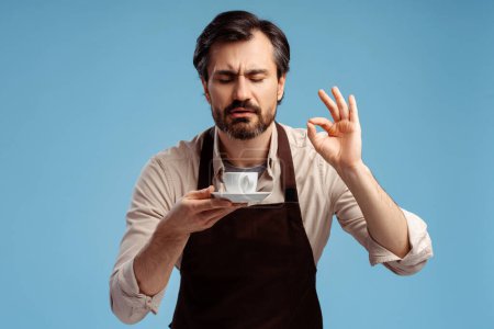 Photo for Young man in apron with closed eyes smelling coffee and showing ok gesture, posing in studio, isolated on blue. Small business startup concept - Royalty Free Image