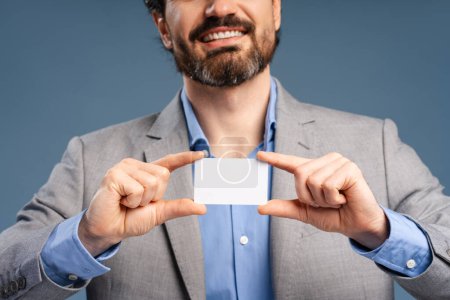 Foto de Hombre de negocios barbudo sonriente sosteniendo la tarjeta en blanco para anunciar aislado sobre fondo azul. De cerca. Logro, carrera, concepto de negocio - Imagen libre de derechos