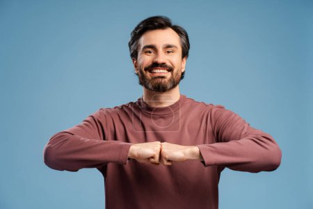 Portrait of smiling young bearded man making fists bump gesture against blue isolated background looking at camera. Advertisement concept