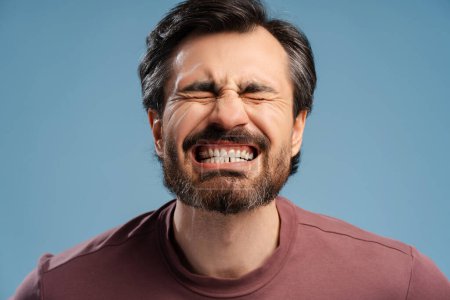 Foto de Retrato de un joven barbudo con los ojos cerrados mostrando su diente aislado sobre fondo azul, posando en el estudio. Dental, ortodoncista, concepto de atención médica - Imagen libre de derechos