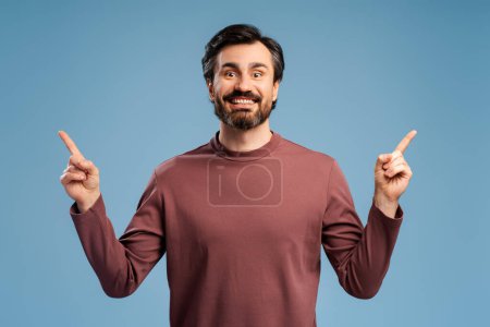 Foto de Portrait of smiling bearded man looking at camera, pointing up by fingers, isolated on blue. Copy space. Shopping, sale concept. Advertisement - Imagen libre de derechos