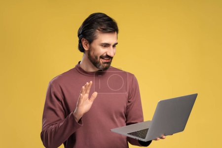 Photo for Smiling positive bearded man using laptop while looking at screen, waving by hand, isolated on yellow background. Online technology concept - Royalty Free Image