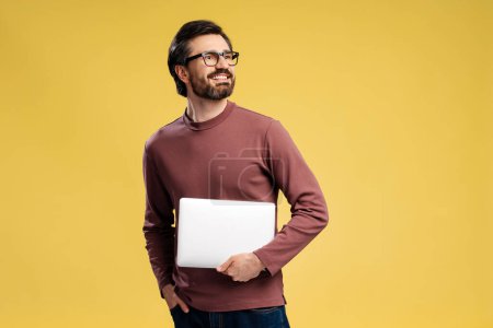 Photo for Portrait of bearded man in glasses holding laptop looking to the side, standing isolated on yellow background. Education, business concept, online technology - Royalty Free Image