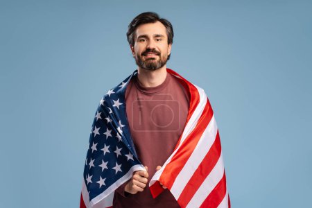 Téléchargez les photos : Portrait de jeune homme heureux et beau avec drapeau américain posant en studio en regardant la caméra, isolé sur bleu. Fête du Souvenir, fête de l'indépendance - en image libre de droit