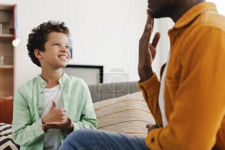 Téléchargez les photos : Petit garçon afro-américain souriant et positif et son père communiquant avec la langue des signes tout en étant assis sur un canapé confortable dans une maison confortable. Concept de soins de santé, relations - en image libre de droit