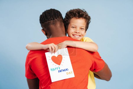 Foto de Atractivo y lindo niño afroamericano sosteniendo la tarjeta de felicitación del día del padre, abrazando a su padre, mirando a la cámara mientras está aislado sobre un fondo azul. Concepto de fiesta, celebración, amor - Imagen libre de derechos