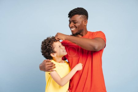 Téléchargez les photos : Heureux souriant père afro-américain et petit fils s'amusent ensemble tout en se tenant isolé sur fond bleu. Concept de parentalité, enfance, publicité - en image libre de droit