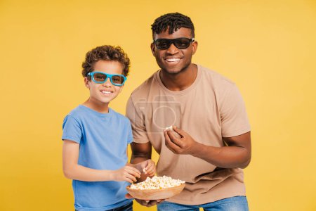 Téléchargez les photos : Un père et un fils afro-américains souriants portant ensemble des lunettes 3D, mangeant du pop-corn, regardant un film ensemble, isolés sur fond jaune. Concept de divertissement - en image libre de droit