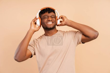 Photo for Attractive african young hipster man wearing wireless headphones listening to music isolated on beige background looking at camera enjoying music, modern technology concept - Royalty Free Image