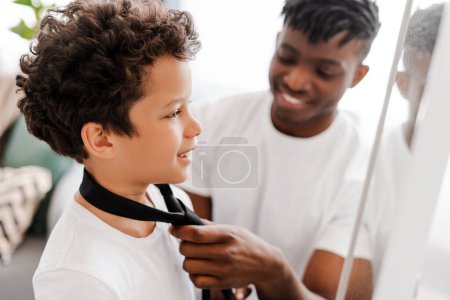 Téléchargez les photos : Beau Afro-Américain, jeune papa habillant son mignon petit fils en cravate, regardant dans un miroir debout dans une chambre confortable à la maison. Concept de passer du temps ensemble, parentalité - en image libre de droit