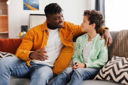 Foto de Familia feliz, padre atractivo y niño pequeño vistiendo ropa colorida hablando mientras está sentado en un cómodo sofá en casa. Concepto de comunicación, paternidad - Imagen libre de derechos