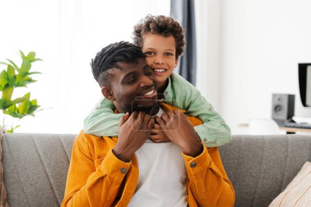 Téléchargez les photos : Heureux jeune père afro-américain et petit fils câlin, assis sur un canapé confortable dans une maison confortable. Concept de parentalité, relations - en image libre de droit