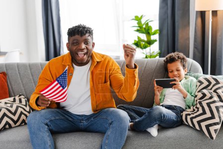 Téléchargez les photos : Happy souriant père afro-américain et petit fils assis sur le canapé à la maison, homme tenant drapeau américain, soutenir, en attente de résultats, petit garçon tenant téléphone mobile, en utilisant l'application mobile - en image libre de droit