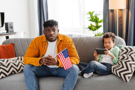 Téléchargez les photos : Homme afro-américain sérieux et petit garçon assis ensemble sur un canapé confortable dans la chambre à la maison, père tenant drapeau américain, fils utilisant smartphone, jeu de jeu. Concept de famille, liberté américaine - en image libre de droit