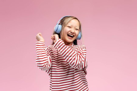 Téléchargez les photos : Heureuse fille asiatique émotionnelle avec écouteurs chantant chanson, écoutant la musique, posant en studio, isolé. Joie et musique. Expression étonnante - en image libre de droit