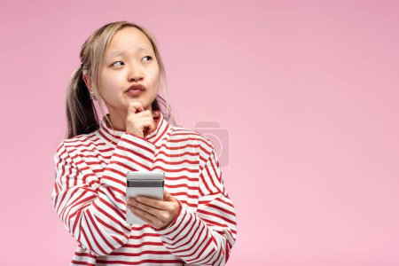 Téléchargez les photos : Adolescent asiatique posant et tenant téléphone portable posant en studio, regardant loin, isolé sur fond rose. Espace de copie. Publicité, concept shopping - en image libre de droit