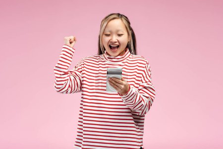 Téléchargez les photos : Fille asiatique émotionnelle posant en studio regardant l'écran du smartphone, tenant la main vers le haut, isolé sur fond rose. Publicité, concept shopping - en image libre de droit