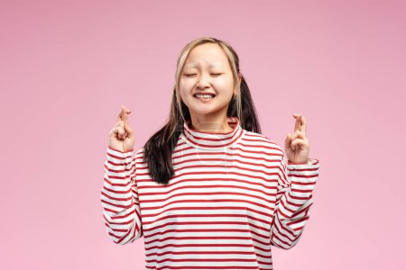 Téléchargez les photos : Jolie fille asiatique posant les yeux fermés tout en gardant les doigts croisés pour jurer isolé sur fond rose. Concept de publicité - en image libre de droit