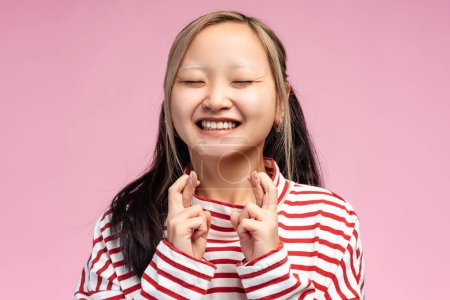 Téléchargez les photos : Joyeux mignon asiatique adolescent fille posant avec les yeux fermés croisant doigt à la caméra, isolé sur fond rose. Ferme là. Concept de publicité - en image libre de droit