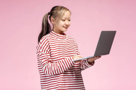 Porträt eines asiatischen Studentenmädchens, das im Studio posiert, Laptop haltend, auf Bildschirm blickend, isoliert auf rosa Hintergrund. Online-Technologie, Bildungskonzept