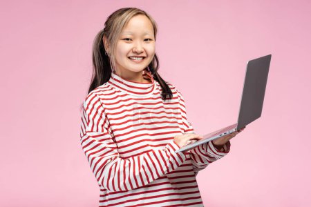 Photo for Happy Asian student girl posing in studio using laptop typing, looking at camera, isolated on pink background. Online technology, education concept - Royalty Free Image