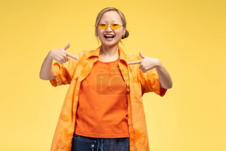 Foto de Retrato de la feliz chica asiática en gafas con estilo mirando a la cámara en el estudio, apuntando a copiar el espacio por los dedos, aislado en amarillo. Concepto de publicidad - Imagen libre de derechos