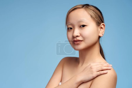 Téléchargez les photos : Portrait de jolie fille asiatique mettant la main sur l'épaule en regardant la caméra, isolé sur bleu, espace de copie. Beauté, corps, concept de soins de la peau - en image libre de droit
