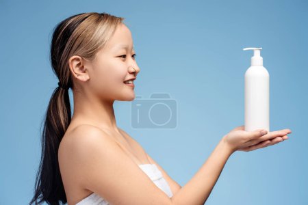 Foto de Vista lateral de la hermosa chica asiática en toalla blanca sosteniendo botella de loción, posando en el estudio, aislado en azul. Concepto de producto de cuidado publicitario. Cuidado de la piel - Imagen libre de derechos