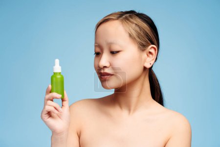 Foto de Retrato de una hermosa chica asiática con hombros desnudos que sostiene el producto de la botella de cosméticos líquidos para la cara limpia, aislado en azul. Cuidado de la piel, belleza, concepto de spa - Imagen libre de derechos