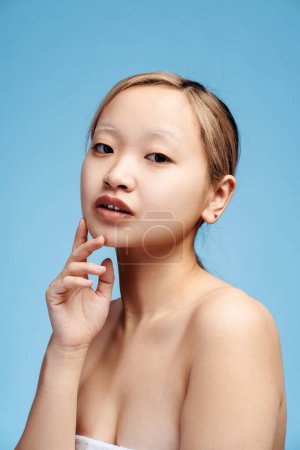 Téléchargez les photos : Portrait de belle fille asiatique debout avec les épaules nues regardant la caméra, tenant la main sur la joue, isolé sur le bleu. Beauté, corps, concept de soins de la peau. Vue verticale - en image libre de droit
