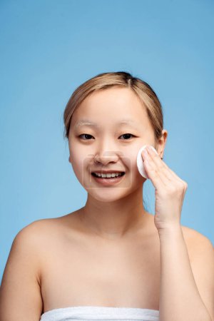 Téléchargez les photos : Portrait de sourire fille asiatique mignonne avec les épaules nues à l'aide de coussinets de coton nettoyer sa peau, isolé sur le bleu. Vue verticale. Soins de la peau, beauté, concept spa - en image libre de droit