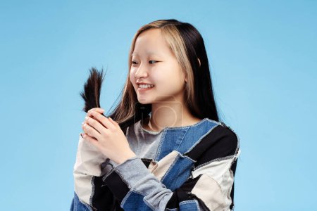 Photo for Portrait of happy Asian girl holding ponytail and looking at the her hair, isolated on blue background. Advertisement, medicine, health care concept - Royalty Free Image