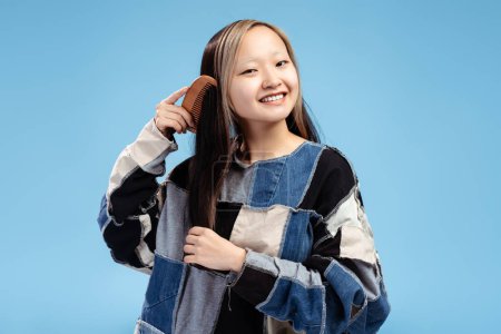Photo for Portrait of smiling pretty Asian girl posing in studio looking at camera while combing her hair, isolated on blue. Advertisement, medicine, health care concept - Royalty Free Image