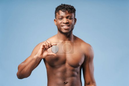 Beau jeune homme afro-américain souriant tenant un stimulateur cardiaque, ICD regardant la caméra, isolé sur fond bleu. Soins de santé, concept de traitement