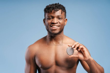 Heureux jeune homme afro-américain souriant tenant un stimulateur cardiaque, ICD regardant la caméra, isolé sur fond bleu. Soins de santé, concept de traitement