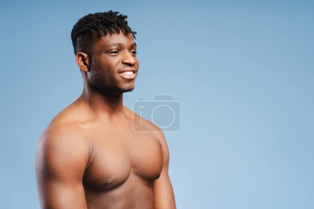 Téléchargez les photos : Souriant homme afro-américain attrayant, athlète nu, torse musculaire regardant loin de l'espace de copie, debout isolé sur fond bleu. Concept de publicité, shopping, mode de vie sain - en image libre de droit