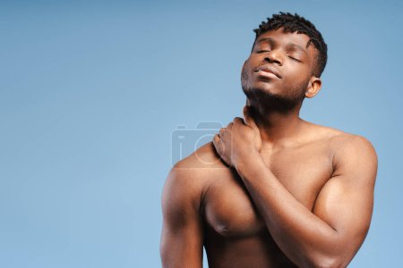 Téléchargez les photos : Pensif, beau, sexy homme afro-américain, mannequin avec torse nu, cou touchant avec les yeux fermés debout isolé sur fond bleu, espace de copie. Concept de publicité - en image libre de droit
