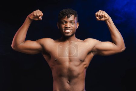 Foto de Sonriente hombre afroamericano guapo, atleta, culturista con el torso desnudo mostrando bíceps en las manos, mirando a la cámara, de pie aislado sobre fondo negro. Concepto de estilo de vida saludable - Imagen libre de derechos