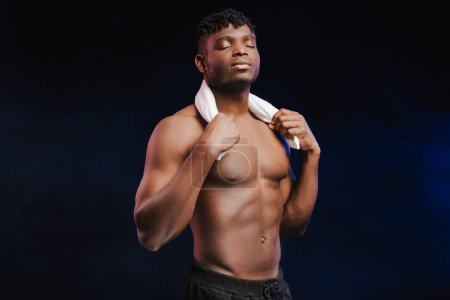 Foto de Guapo hombre afroamericano serio, atleta, culturista con torso desnudo sosteniendo la toalla con los ojos cerrados de pie aislado sobre fondo negro. Concepto de entrenamiento, estilo de vida saludable - Imagen libre de derechos