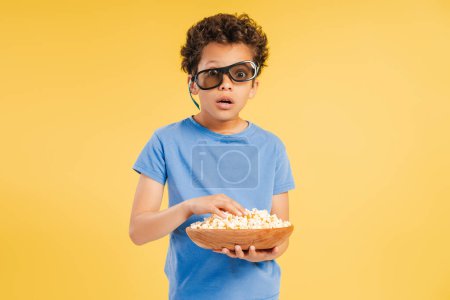 Téléchargez les photos : Choqué excité mignon garçon portant des lunettes 3D manger pop-corn regarder le film en regardant la caméra debout isolé sur fond jaune. Concept de divertissement - en image libre de droit