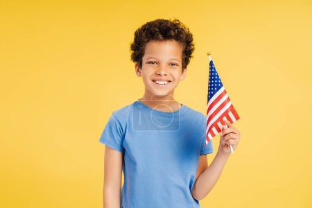 Téléchargez les photos : Petit garçon afro-américain souriant, patriote tenant le drapeau américain regardant la caméra debout isolé sur fond jaune. Concept des élections américaines, fête de l'indépendance, 4 juillet - en image libre de droit