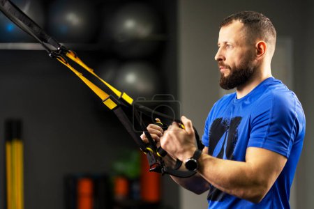 Serious, strong man, athlete holding loops of trx equipment is training in the gym. Concept of functional training, sport