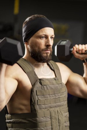 Photo for Portrait of attractive, young, bearded man wearing weight plate, holding dumbbells, doing exercises, looking away, working out in modern gym. Concept of sports, training, healthy lifestyle - Royalty Free Image