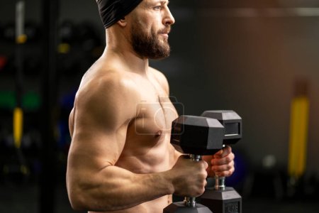 Téléchargez les photos : Handsome, muscular, bearded man with naked torso holding dumbbells, training in gym and looking away. Concept of sport, healthy lifestyle - en image libre de droit