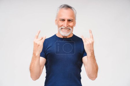Glücklicher gutaussehender grauhaariger Mann, der T-Shirt über weißem Hintergrund trägt und Rocksymbol mit erhobenen Händen in die Kamera schaut. Musikkonzept