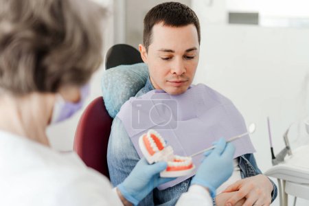 Senior attraktive Zahnärztin mit Kiefer, zeigt geduldigen, gutaussehenden jungen Mann, erklärt etwas, arbeitet in einer modernen Zahnklinik. Konzept der Gesundheitsfürsorge, Zahnpflege, Behandlung